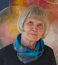 Gudrun Bertsche mit einem lächelnden Gesicht, blauem Pullover, grün-blauem Seidenschal und grauen halblangen Haaren steht vor einem ihrer farbenfrohen Kunstwerken. 