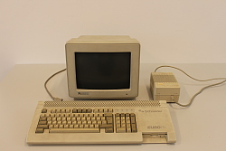 Es ist ein alter Conmputer aus den 1980er Jahren zu sehen. Der Monitor ist klein und tief. Davor liegt eine Tastatur, abgenutzt und mit hohen Tasten. Rechts neben dem Monitor findet sich ein rechteckiges Netzteil.