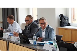 Vier Männer an einem langen Tisch mit Mikrofonen