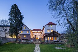 Beleuchtetes historisches Gebäude mit mehreren Giebeln und rotem Dach, umgeben von Bäumen und einem Weg im Vordergrund.