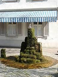 Brunnen aus aufgetürmten Steinen, die von Moos bewachsen sind, steht auf Platz aus Kopfsteinpflaster, weiße Hausfassade mit blau-weiß gestreifter Markise im Hintergrund