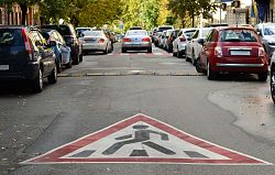 Zu sehen ist eine Straße in einer Stadt, auf der ein Auto fährt, das von hinten zu sehen ist. Rechts und links sieht man Häuser und davor parkende Autos. Im Vordergrund ist auf dem Asphalt das Piktogramm eines Fußgängerüberwegs aufgemalt. Die Straße ist von grünen Laubbäumen gesäumt.