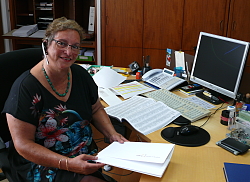 Petra Bredemeyer sitzt am Schreibtisch, vor ihr ein PC und hält eine weiße Lahr-Mappe in der Hand. Sie trägt ein Headset und lächelt von unten in die Kamera. 