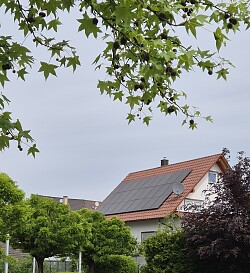 Einfamilienhaus mit Solarpanelen auf dem roten Dach, umgeben von grünen Bäumen unter bewölktem Himmel.