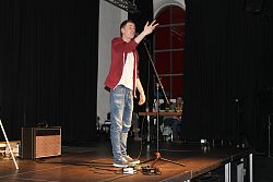 Großaufnahme von Andreas Rebholz während seines Auftritts auf der Schlachthof-Bühne. Er trägt blaue Jeans, ein weißes T-Shirt und eine rote geöffnete Sweatjacke.