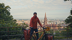 Der Filmemacher steht mit seinem Fahrrad auf einem Berg, im Hintergrund die Silhouette von Freiburg. 