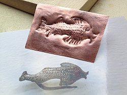 Das Bild zeigt eine kupferfarbene Metallplatte, in die ein Fisch punziert wurde und das Gegenstück dazu.