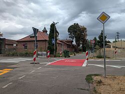 Straßenkreuzung mit rotem Radweg, mehreren Verkehrszeichen und Backsteingebäuden im Hintergrund.