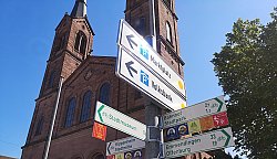 Radwegweiser an der Stiftskirche Lahr: An einer Metallstange sind verschiedene Wegweiser angebracht. Zwei zeigen Parkhäuser an und vier Wegweiser zeigen Radwege zu Zielen in Lahr und in umliegende Gemeinden. Im Hintergrund die Türme der Stiftskirche vor blauem Himmel