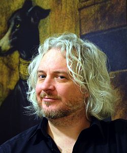 Portraitfoto des Autors Ralf H. Dorweiler. Er hat hellblondes, mittellanges, gewelltes Haar, trägt einen Bart an Oberlippe, Kinn und Wangen. Bekleidet ist er mit einem dunkelblauen Hemd, dessen obere Knöpfe geöffnet sind. Im Hintergrund ist das Gemälde eines Hundes zu sehen.