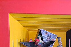 Zu sehen ist eine offene Holzbox, die mit den Farben der Deutschlandfahne bemalt wurde. Im Inneren sind verschiedene Objekte verarbeitet: von einer sternförmigen Schachtel über einen Bollenhut bis hin zu Stiften und einer Farbtube sowie einer selbstgemachten Corona-Maske.