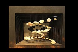 Das Raumwunder, eine Holzbox mit einer großen Öffnung vorne, ist schwarz. In dem freien Raum der Box hängen verschieden farbige und große Kieselsteine an Perlonfände von der Decke der Box. Die Kieselsteine sind in einer Spirale angeordnet.