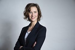 Portrait einer lächelnden Frau mit blauen Augen und halblangen braunen welligen Haaren. Sie trägt ein dunkelblaues Sakko, weißes T-Shirt und hat die Arme vor der Brust verschränkt.