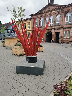 Zu sehen ist Reinhard Sigles Werk &#34;EinschlagAuswux&#34;, welches sich auf dem Rathausplatz in Lahr befindet. Geborstenes Holz in roter Farbe richtet sich gen Himmel.