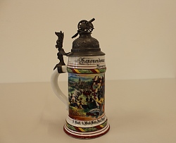 Das Bild zeigt einen weißen Krug mit Henkel und aufwändigem Metalldeckel, auf dem eine Kanone steht. Auf dem Krug ist Lahr abgebildet sowie Soldaten.