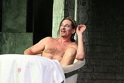 Der Schauspieler Max Tidof liegt oberkörperfrei in einer Badewanne auf der Bühne.