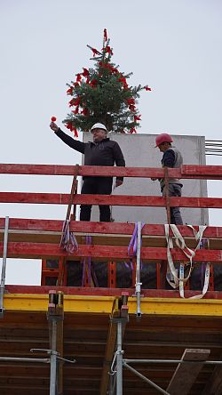 Zwei Männer mit Bauhelmen stehen auf einem Gerüst vor einem mit roten Bändern geschmückten Tannenbaum, einer von beiden hält ein Glas Rotwein in die Höhe