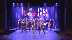 Das Foto zeigt die Band während einem Auftritt auf der Bühne. Im Vordergrund der Sänger mit den Tänzerinnen. Im Hintergrund die Band mit bzw. an ihren Instrumenten.