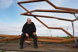 Der Künstler Robert Schad sitzt auf einem Anhänger links neben seinem Kunstwerk. Dieses besteht aus zersägten und zusammengeschweißten Baustahlstäben in rostbraun. Die Skulptur ist größer als der Künstler und liegt seitwärts auf der Ladefläche. Der Himmel im Hintergrund ist blau. Robert Schad trägt schwarze Kleidung und hat kurze weiße Haare. Er blickt auf sein Kunstwerk.