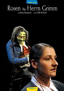 Im Vordergrund zu sehen ist Katja Baumann. Sie hat dunkles glattes Haar im Nacken zu einem Dutt gebunden und trägt eine helle Bluse und eine graue Jacke. Hinter ihr sieht man eine große Marionette auf einem Stuhl sitzen.