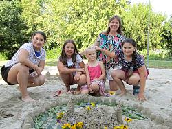 Vier Mädchen und eine erwachsene Frau hocken lachen neben einer Sandburg im Sand. Sie haben sichtlich Spaß beim Sanburgenbauen und haben ihr Werk mit bunten Blümchen verziert.