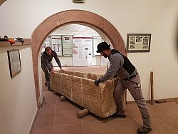 Zwei Männer schieben einen großen Steinsarkophag durch eine bogenförmige Wandöffnung im Untergeschoss der Villa Jamm.