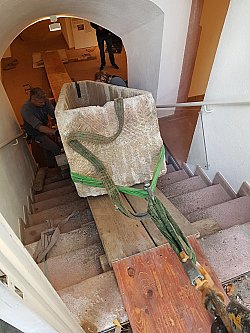 Zu sehen ist der Transport eines Steinsarkophags aus der Villa Jamm hinaus über eine steile Treppe nach oben. Auf die Treppenstufen wurden Holzplanken gelegt. Der Sarkophag ist an Gurten befestigt.