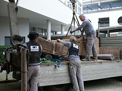 Auf dem Bild sieht man drei Männer der Firma Nuvolin wie sie den Sarkophag mit einem Kran von einem Lastwagen abladen.