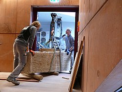 Das Bild zeigt den Sarkophag en einem Kran hängend. Drei Arbeiter des Steinmetzbetriebs bewegen den großen Stein zu einer Tür hinein in die Tonofenfabrik.