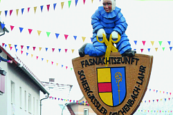 Das Bild zeigt die Schergässler in Aktion beim Fasentumzug. Vornweg marschiert ein Schergässler im blauen Filz-Häs mit Holzmaske, weißen Handschuhen und weißer Ganzkopfmütze mit zwei Hörnern, der ein Schild mit dem Reichenbacher Wappen und ringsherum der Aufschrift Fasnachtszunft Schgässler Lahr-Reichenbach trägt. Oben auf dem Schild sitzt eine Schergässler-Figur im Häs aus Holz. Hinter dem Schild-Träger laufen zahlreiche weitere Schergässler im blauen Filz-Häs mit Holzmaske, weißer Ganzkopfmütze mit Hörner, weißen Handschuhen und gelben Scheren.