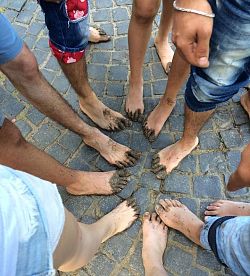 Das Bild zeigt die Beine mehrerer Personen, die im Kreis stehen. Sie haben kurze Hosen an oder die Hosenbeine hochgekrempel und stellen ihre nackten, mit Schlamm beschmierten Füße im Kreis zusammen. 