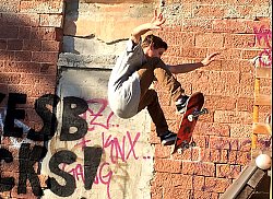 Zu sehen ist ein junger Mann, der in einer Halfpipe mit dem Skateboard einen Sprung ausführt. Im Hintergrund ist eine rote Ziegelwand mit Grafitti zu sehen.