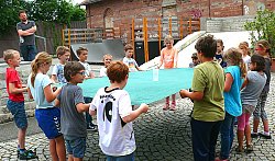 Teamtraining mit Heiko Heimburger: Im Hof beim Schlachthof steht Heiko Heimburger und beobachtet eine Schülergruppe, die gemeinsam einen Becher mit Wasser auf einem großen Tuch balancieren muss. Die Schülerinnen und Schüler halten jeder ein Stück vom Tuch fest, gemeinsam müssen sie darauf achten, dass der gefüllt Becherm der in der Mitte des Tuches steht, nicht umfällt.