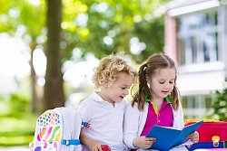 Zwei Schulkinder, ein Junge und ein Mädchen sitzen auf einer Bank. Beide sind im Alter der ersten Klasse oder Vorschule. Das Mädchen hlt ein blaues Buch in den Händen, aus dem sie vorzulesen scheint. Sie trägt Zöpfe, ein pinkfarbesnes Polo-Shirt mit grünem Kragen, einen weißen Rock und eine weiße Jacke. Der Junge neben ihr hat blondes lockiges Haar und trägt ein weißes Polo-Shirt und eine blaue Hose. In einer Hand hält er einen Apfel. Neben den Kindern stehen bunte Schulrucksäcke auf der Bank. Und neben dem Mädchen liegt ein Stapel bunter Bücher.