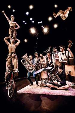Band mit Musikinstrumenten spielt Musik, ein Mann macht gerade einen Salto in der Luft, ein Mann fährt Einrad und hat dabei einen weiteren Mann auf seinen Schultern sitzen, auf dessen Schultern eine Frau steht.