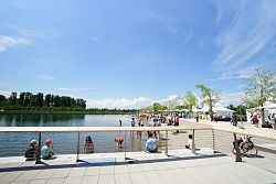 Das Bild zeigt einen Sommertag am Stegmattensee in Lahr während der Landesgartenschau 2018. Man sieht die Uferpromenade mit weißen Zelten und zahlreichen Besuchern. Am Seeufer sind viele Kinder, die im Sand spielen oder Eis essen. Im Hintergrund das gegenüberliegende Ufer mit viel Grün und Bäumen.
