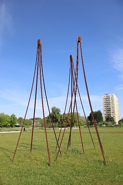 3 Skulpturen aus Cortenstahl, bestehend jeweils aus 4 Stahlstäben, die spitz zusammenlaufen und sich in der Spitze treffen, wo ein winzig kleines Vogelhäuschen angebracht ist. Wiese und Hochhaus im Hintergrund.