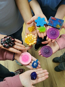 Mehrere Hände halten bunte, in verschiedenen Formen gegossene Seifenstücke wie Schneeflocke, Biene, Schildkröte und Blume.