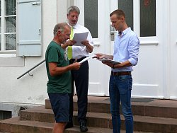 Martin Stehr überreicht einem Herrn in einem grünen T-Shirt und kurzen Hosen vor dem Rathaus eine Urkunde für die erfolgreiche Teilnahme am Stadtradeln 2018. Bürgermeister Tilman Petters steht daneben und liest aus einer Liste vor.