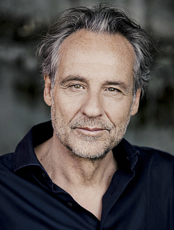 Portraitbild eines Mannes mit schwarz-grauen Haaren, braunen Augen und Dreitagebart. Er trägt ein schwarzes geöffnetes Polohemd.