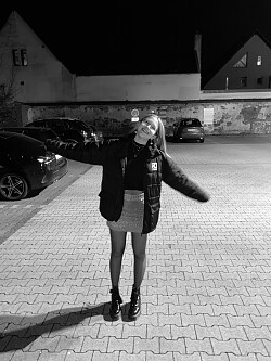 Das Bild wurde in schwarz-weiß aufgenommen. Die Künstlerin steht nachts auf einem Parkplatz und lacht die Kamera in einer lockeren, beschwingten Bewegung an.