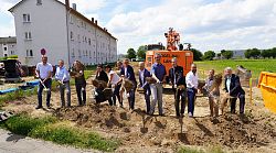 13 Männer und Frauen stehen nebeneinandern und stechen mit Spaten in die Erde, im Hintergrund ein Wohnhaus und blauer Himmel.