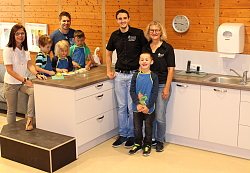 Sabine Grothe ( Leiterin), Marius Schlager ( Erzieher), Adrian Mussler und Susanne Mussler ( eschäftsführerin)von Mussler Küchen und Kinder der Kita Am Schießrain stehen vor der neuen weißen Küche, teilweise auf Podesten. &#13;&#10;
