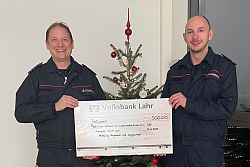 Zwei Feuerwehrmänner vor einem Weihnachtsbaum halten einen großen Spendenscheck