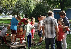 Zu sehen ist eine Gruppe Kinder mit drei Erwachsenen. Sie stehen auf einer Wiese, umringt von Bäumen, an einer Obstpresse und pressen Apfelsaft. Im Hintergrund steht das Spielmobil, ein Traktor mit blauem Bauwagen, auf dem in bunter Schrift &#34;Spielmobil&#34; steht.