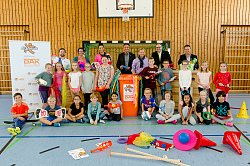 Bei der Auftaktveranstaltung zu fit4future haben sich die Verantwortlichen mit den Schülern zu einem Gruppenbild zusammengestellt. Davor liegen Spielgeräte, die Kinder halten jeweils ein Spielgerät in der Hand. In der Mitte steht die orangene Spieltonne. 