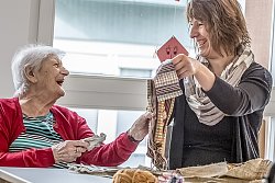 Zu sehen ist eine Seniorin, die an einem Tisch sitzt, auf dem Stoffe und Wollreste liegen. Sie schaut lachend zu einer jüngeren Frau, die neben ihr am Tisch steht und ein Stück karierten Stoff hochhält. Sie schaut die ältere Frau ebenfalls lachend an.