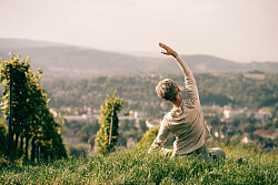 Das Foto zeigt eine Dame die Yoga inmitten der Reben auf dem Schutterlindenberg macht. Im Hintergrund sind Häuser in der Ferne und die grünen Wälder der Umgebung zu sehen. 