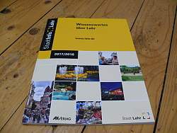 Zu sehen ist die StadtInfo Broschüre Lahr 2017/18 auf einem Holzboden liegend, schräg von vorn fotografiert. Die Broschüre hat auf der Titelseite mehrere quadratische Fotos und ist in der Grundfarbe Gelb gehalten.