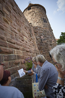 Stadtführung in der Stadt Lahr. Eine Gruppe steht am Lahrer Storchenturm und lauscht den Ausführungen einer Stadtführerin. Sie zeigt ein Bild der alten Anlage der Wasserburg. 